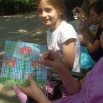 Ein Mädchen sitzt im Freien und lächelt in die Kamera, während eine andere Person ein Stück Plastikleinwand mit einem farbenfrohen Stickereimotiv in der Hand hält.