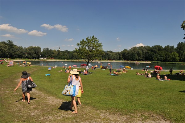 Menschen entspannen sich auf einer Wiese an einem See unter einem klaren Himmel, einige sonnen sich, schwimmen und tragen Taschen zum Wasser. Der Hintergrund ist von Bäumen gesäumt.