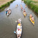 Eine Gruppe von Menschen paddelt an einem bewölkten Tag mit Kanus einen schmalen Fluss entlang, der von Grasfeldern und Schilf gesäumt ist.