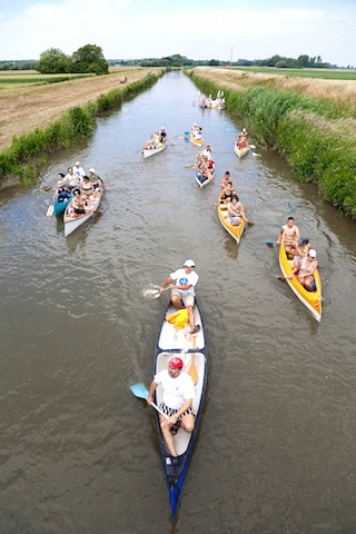 Eine Gruppe von Menschen paddelt an einem bewölkten Tag mit Kanus einen schmalen Fluss entlang, der von Grasfeldern und Schilf gesäumt ist.