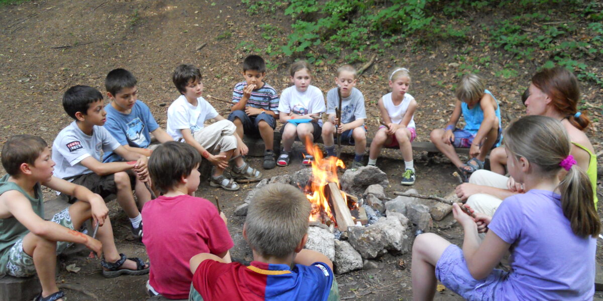 Eine Gruppe von Kindern und ein Erwachsener sitzen auf Bänken im Kreis um ein Lagerfeuer in einem Waldstück.