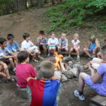 Eine Gruppe von Kindern und ein Erwachsener sitzen auf Bänken im Kreis um ein Lagerfeuer in einem Waldstück.