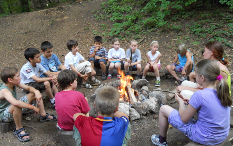 Eine Gruppe von Kindern und ein Erwachsener sitzen auf Bänken im Kreis um ein Lagerfeuer in einem Waldstück.