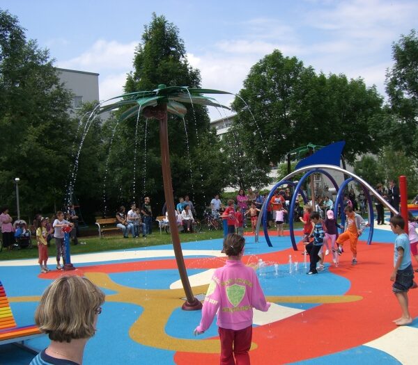 Kinder spielen in einem farbenfrohen Planschbecken mit Wasserspielen in Form von Palmen, während Erwachsene von Bänken im Hintergrund zusehen.