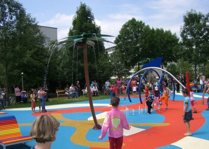 Kinder spielen in einem farbenfrohen Planschbecken mit Wasserspielen in Form von Palmen, während Erwachsene von Bänken im Hintergrund zusehen.
