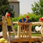 Drei Kinder mit roten Halstüchern spielen auf einem hölzernen Spielplatz im Freien, wobei ein Kind ein blaues Rad dreht; im Hintergrund sind Bäume und ein Gebäude zu sehen.