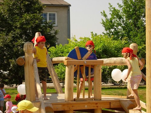Drei Kinder mit roten Halstüchern spielen auf einem hölzernen Spielplatz im Freien, wobei ein Kind ein blaues Rad dreht; im Hintergrund sind Bäume und ein Gebäude zu sehen.