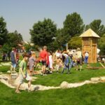 An einem sonnigen Tag versammeln sich Kinder und Erwachsene auf einer Spielwiese mit einem Holzspielgerät und Bäumen im Hintergrund.