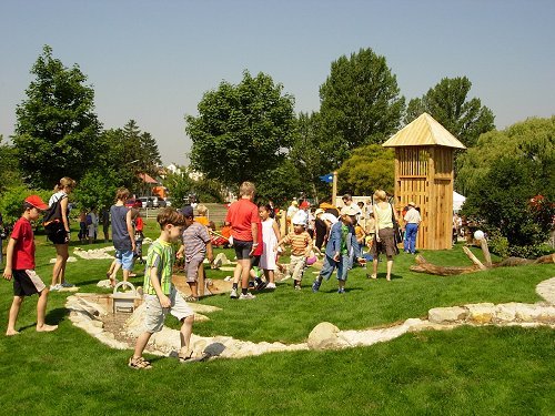 An einem sonnigen Tag versammeln sich Kinder und Erwachsene auf einer Spielwiese mit einem Holzspielgerät und Bäumen im Hintergrund.