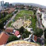 Luftaufnahme eines großen Stadtparks mit einem Spielplatz, Spazierwegen und Menschenansammlungen, umgeben von städtischen Gebäuden und Grünflächen.