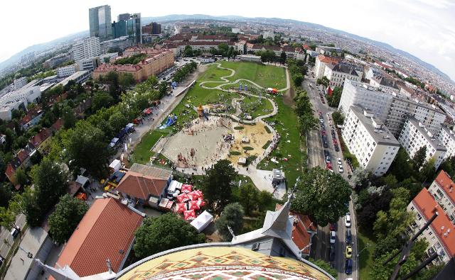 Luftaufnahme eines großen Stadtparks mit einem Spielplatz, Spazierwegen und Menschenansammlungen, umgeben von städtischen Gebäuden und Grünflächen.