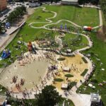 Luftaufnahme eines belebten Parks mit einem großen Spielplatz, einem Planschbecken, Spazierwegen, Rasenflächen und Menschen, die an einem sonnigen Tag verschiedene Aktivitäten genießen.
