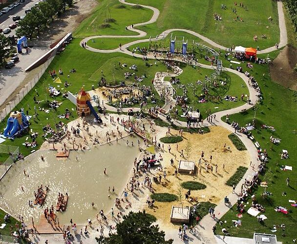 Luftaufnahme eines belebten Parks mit einem großen Spielplatz, einem Planschbecken, Spazierwegen, Rasenflächen und Menschen, die an einem sonnigen Tag verschiedene Aktivitäten genießen.