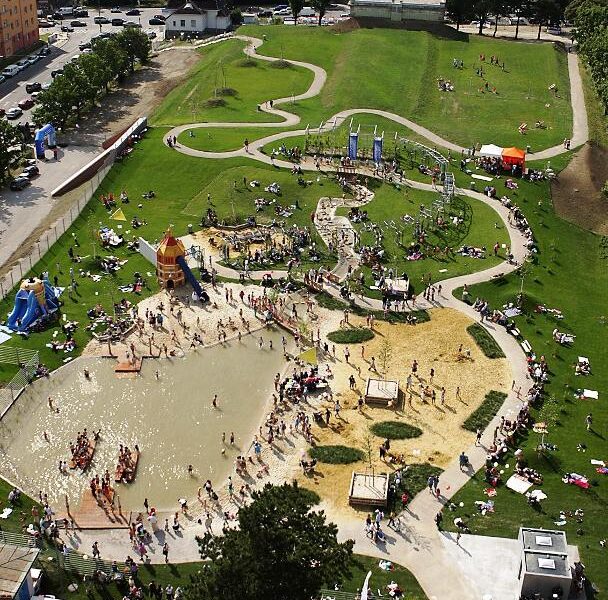 Luftaufnahme eines belebten Parks mit einem großen Spielplatz, einem Planschbecken, Spazierwegen, Rasenflächen und Menschen, die an einem sonnigen Tag verschiedene Aktivitäten genießen.