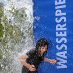 Ein Kind läuft durch einen Wasservorhang auf einem Spielplatz im Freien, neben einem blauen Schild mit der Aufschrift "WASSERSPIELPLATZ".