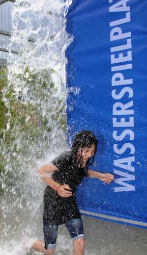 Ein Kind läuft durch einen Wasservorhang auf einem Spielplatz im Freien, neben einem blauen Schild mit der Aufschrift "WASSERSPIELPLATZ".