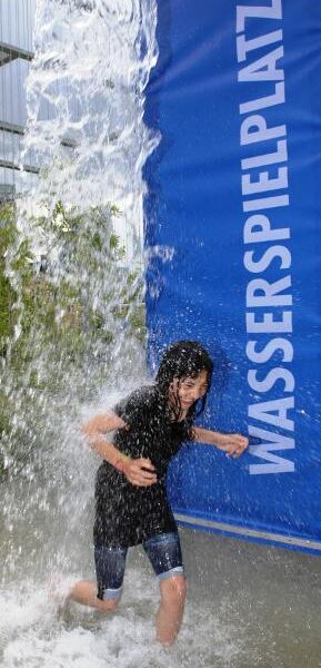 Ein Kind läuft durch einen Wasservorhang auf einem Spielplatz im Freien, neben einem blauen Schild mit der Aufschrift "WASSERSPIELPLATZ".