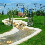 Ein seichter Bach fließt durch einen grasbewachsenen Park, der von Felsen und Metallbögen gesäumt ist, mit einer Holzbrücke und einer Bank im Hintergrund.