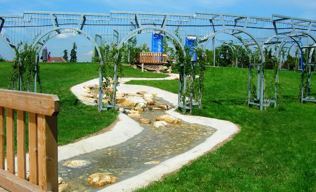 Ein seichter Bach fließt durch einen grasbewachsenen Park, der von Felsen und Metallbögen gesäumt ist, mit einer Holzbrücke und einer Bank im Hintergrund.