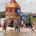 Kinder und Erwachsene versammeln sich an einem sonnigen Tag um ein hölzernes Spielgerät mit einer blauen Rutsche in der Nähe eines flachen Wasserbereichs und spielen.