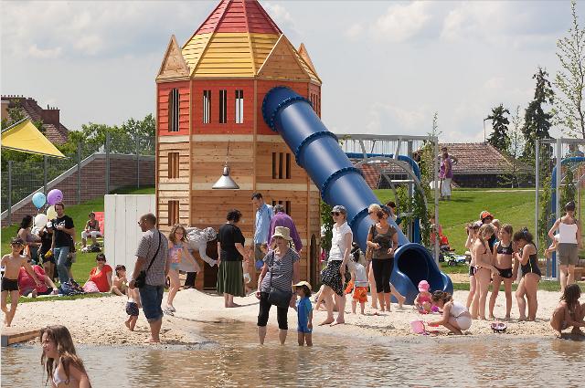 Kinder und Erwachsene versammeln sich an einem sonnigen Tag um ein hölzernes Spielgerät mit einer blauen Rutsche in der Nähe eines flachen Wasserbereichs und spielen.