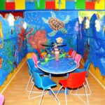 Farbenfroher Kinderpartyraum mit Unterwasserthema, mit Meeresmalereien an den Wänden, einem runden Tisch, der für eine Party gedeckt ist, und leuchtend roten, blauen und rosa Stühlen.