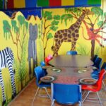 Kinderspielzimmer mit einem Wandgemälde zum Thema Dschungel, das ein Zebra, einen Elefanten, eine Giraffe und einen Affen zeigt, und einem Tisch, der von bunten Stühlen umgeben ist.