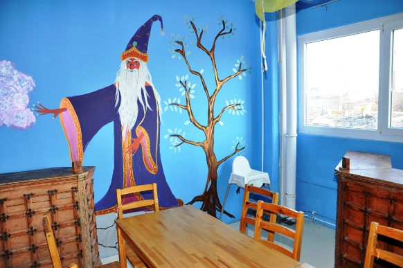 Ein Klassenzimmer mit einem Wandgemälde von einem Zauberer und einem Baum an einer blauen Wand, Holztischen und -stühlen, Truhen, einem Hochstuhl und einem Fenster, das Tageslicht hereinlässt.