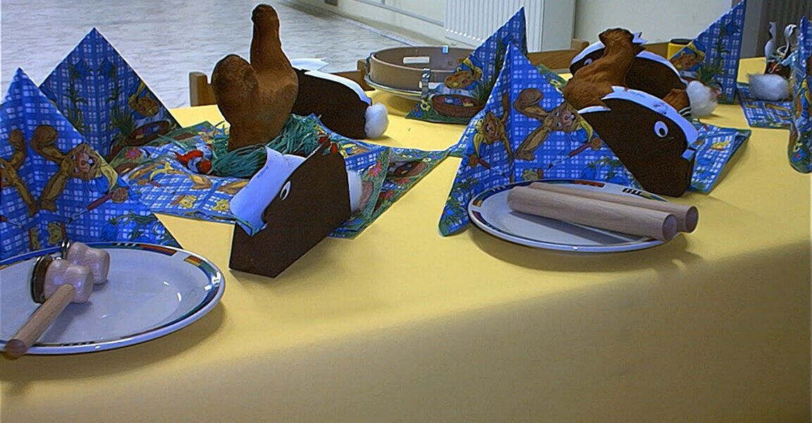 Ein gedeckter Tisch mit gelber Tischdecke, Tellern, Holzbesteck, gefalteten Servietten und Hähnchenkeulen aus Plüsch. Im Hintergrund stehen grüne Stühle und ein weißer Heizkörper.