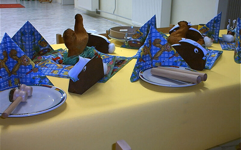 Ein gedeckter Tisch mit gelber Tischdecke, Tellern, Holzbesteck, gefalteten Servietten und Hähnchenkeulen aus Plüsch. Im Hintergrund stehen grüne Stühle und ein weißer Heizkörper.