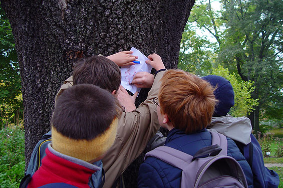 Vier Kinder mit Rucksäcken drängen sich um einen Baum und schreiben oder zeichnen mit einem Marker auf ein Stück Papier, das im Freien an den Baumstamm gedrückt wird.