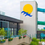 Eine große Wasserrutsche im Freien windet sich um einen Turm mit einem Sonnen- und Wellenlogo neben einem Gebäude mit Topfpflanzen und Geländern.