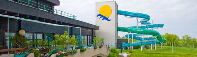 Eine große Wasserrutsche im Freien windet sich um einen Turm mit einem Sonnen- und Wellenlogo neben einem Gebäude mit Topfpflanzen und Geländern.