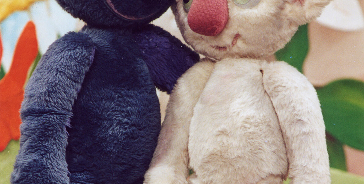 Zwei plüschige Koala-Puppen, eine blaue und eine weiße, stehen nebeneinander vor einem bunten, illustrierten Hintergrund.
