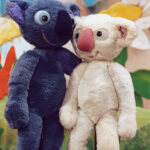 Zwei plüschige Koala-Puppen, eine blaue und eine weiße, stehen nebeneinander vor einem bunten, illustrierten Hintergrund.