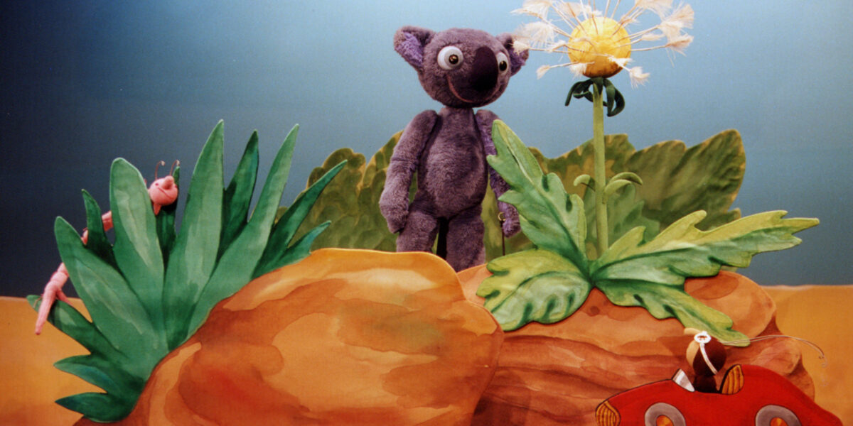 Ein lilafarbener Plüschbär steht neben einer Pusteblume zwischen Steinen, mit einem Spielzeugwurm auf einem Blatt und einer Maus in einem roten Spielzeugauto im Vordergrund.