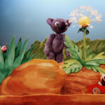 Ein lilafarbener Plüschbär steht neben einer Pusteblume zwischen Steinen, mit einem Spielzeugwurm auf einem Blatt und einer Maus in einem roten Spielzeugauto im Vordergrund.