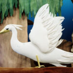 Ein weißer Puppenvogel mit erhobenen Flügeln steht auf einem Ast vor einer Kulisse mit Bäumen; im Hintergrund ist eine blaue teddybärähnliche Puppe zu sehen.