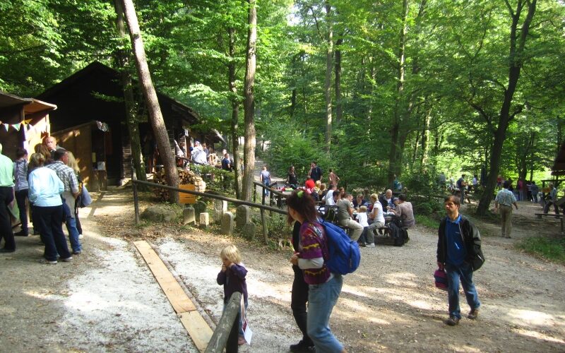 Die Menschen versammeln sich an Picknicktischen und spazieren an einem sonnigen Tag durch ein Waldgebiet in der Nähe von Holzhütten unter Bäumen.