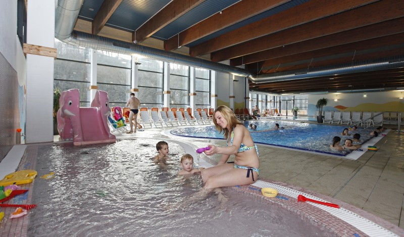 Ein Hallenbad mit Kindern und Erwachsenen; eine Frau sitzt mit zwei kleinen Kindern am Beckenrand, während andere in der Nähe schwimmen und spielen.