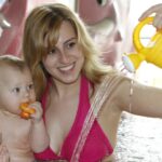 Eine Frau in einem rosa Bikini hält ein Baby in einem Pool, während sie Wasser aus einer gelben Gießkanne gießt; das Baby hält ein Spielzeug und schaut ins Wasser.