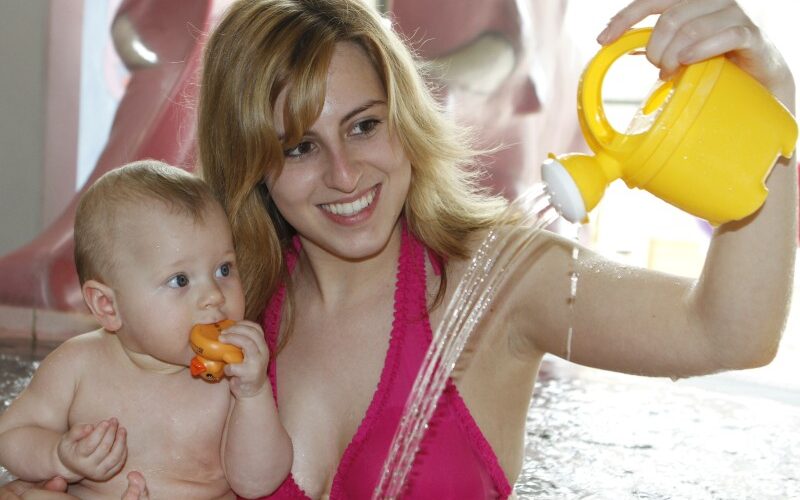 Eine Frau in einem rosa Bikini hält ein Baby in einem Pool, während sie Wasser aus einer gelben Gießkanne gießt; das Baby hält ein Spielzeug und schaut ins Wasser.