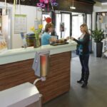 Eine Frau steht an einer Rezeption und spricht mit einer Empfangsdame in einer hell erleuchteten Lobby mit Stühlen, Broschüren und Dekorationen.