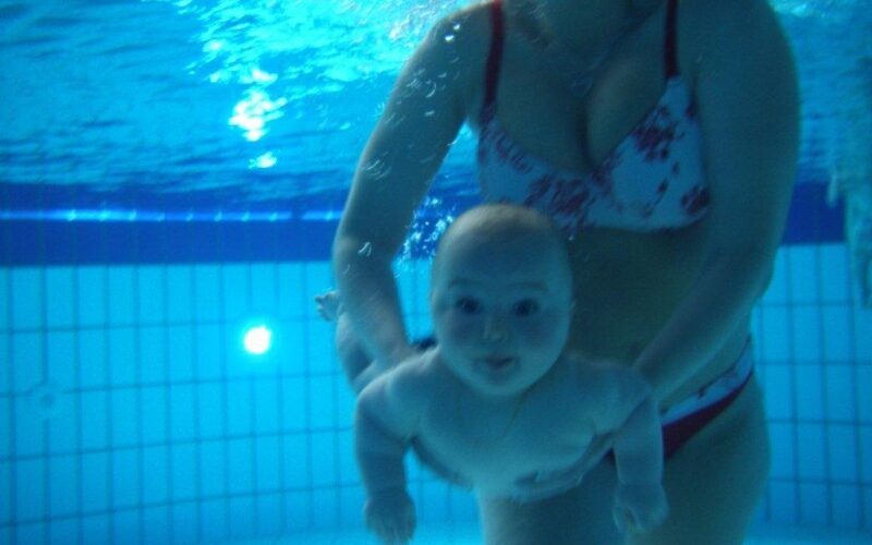 Ein Erwachsener im Badeanzug hält ein Baby unter Wasser in einem Schwimmbad. Beide sind untergetaucht, und im Hintergrund sind die Beckenfliesen zu sehen.