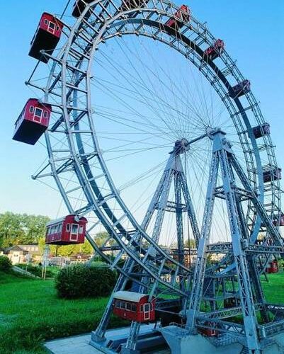 Ein großes Riesenrad mit roten Fahrgastkabinen steht auf einer Wiese unter einem strahlend blauen Himmel.