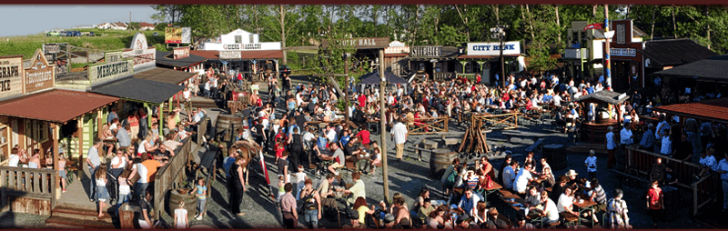 Eine große Menschenmenge versammelt sich bei sonnigem Wetter in einer Stadt im Freien, die dem alten Westen nachempfunden ist, mit Holzgebäuden und verschiedenen Schildern.