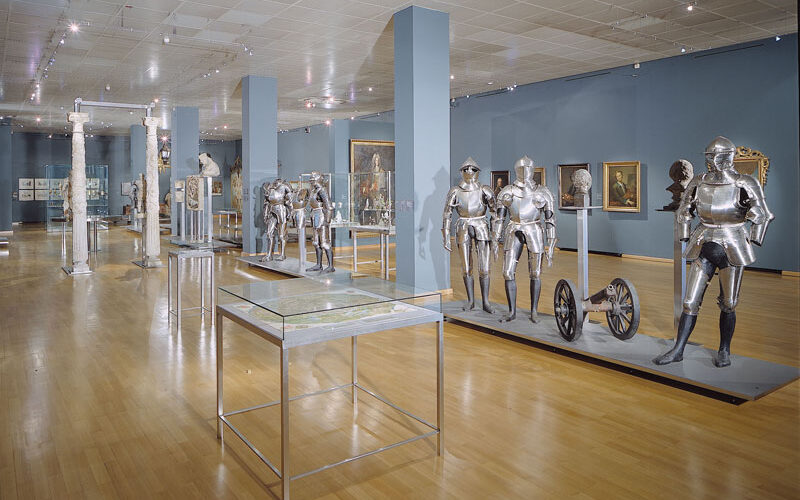 Museumsgalerie mit Exponaten von mittelalterlichen Rüstungen, Helmen und Waffen, die in Vitrinen und auf Ständern ausgestellt sind; blaue Wände und polierter Holzboden.