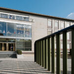 Außenansicht des Wien Museums Karlsplatz mit dem Haupteingang, großen Fenstern und einem Geländer im Vordergrund bei teilweise bewölktem Himmel.