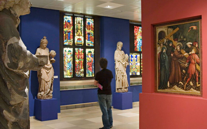 Eine Person steht in einer Museumsgalerie mit Statuen, Buntglasfenstern und einem Gemälde, das eine religiöse Szene an der Wand darstellt.