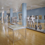 Museumsgalerie mit Glasvitrinen, mittelalterlichen Rüstungen, Helmen und historischen Artefakten, die auf Ständern vor blauen Wänden und einem polierten Holzfußboden aufgestellt sind.